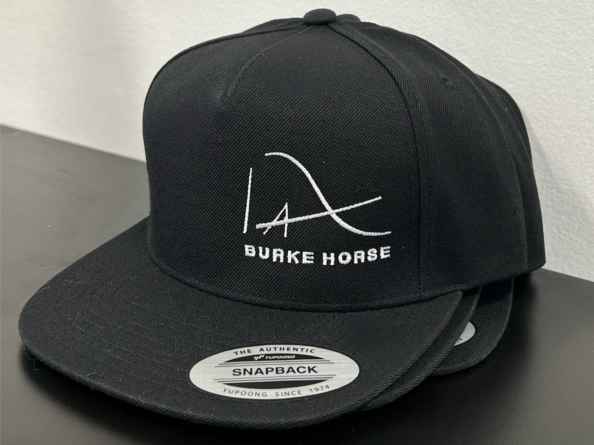 A black snapback hat with Burke Horse logomark and text in white on the front.